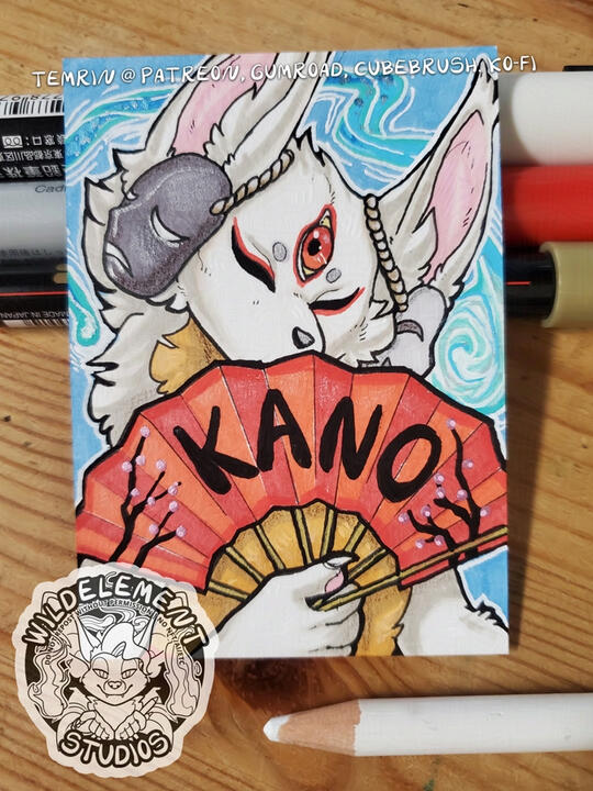 a photo of a 2x3 inch badge on a wooden surface, with some pens and pencils around it. The badge has a blue background with a magical whisp around the edges, with a white kitsune character in the center, holding a red fan that reads "KANO" and the shapes o